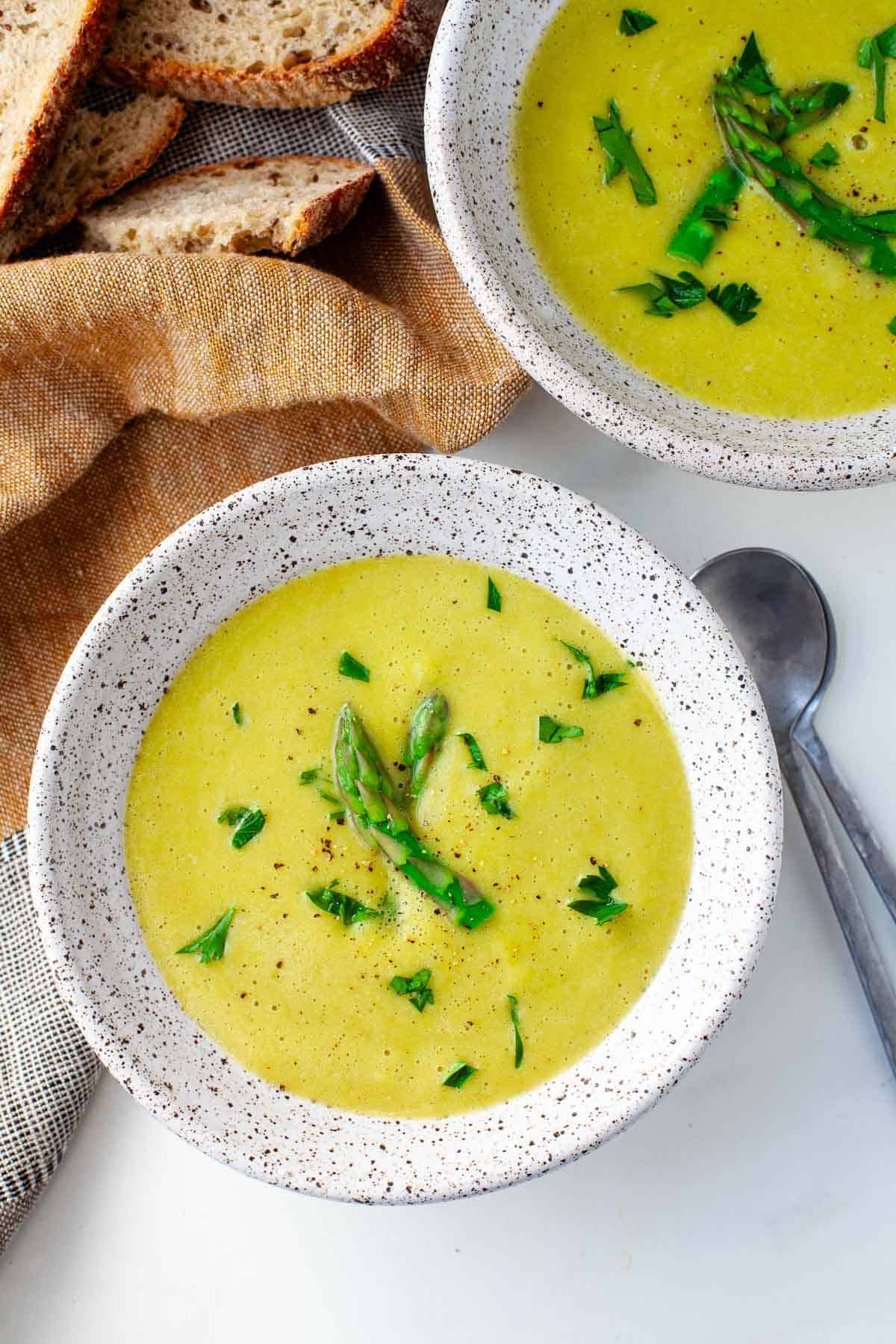 vegan asparagus soup in two white speckled bowls beside two silver spoons, a kitchen towel, and artisan bread