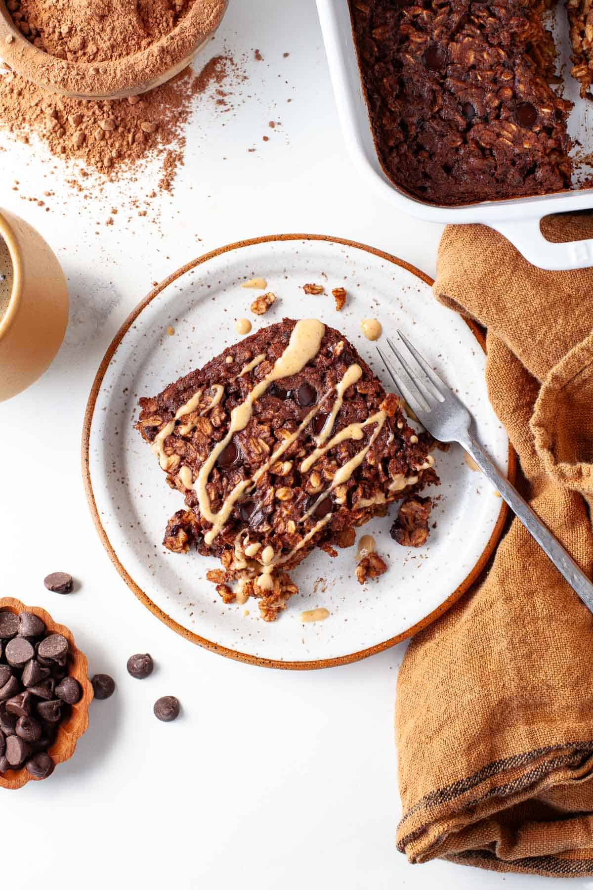 chocolate baked oatmeal on plate surrounded by kitchen towel, cocoa powder, chocolate chips, and baking dish.